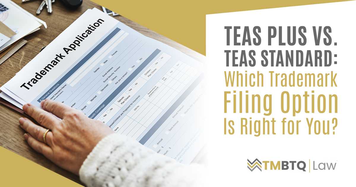 A hand rests on a printed form labeled 'Trademark Application' on a wooden desk, representing the decision between TEAS Plus and TEAS Standard filing options with the USPTO.